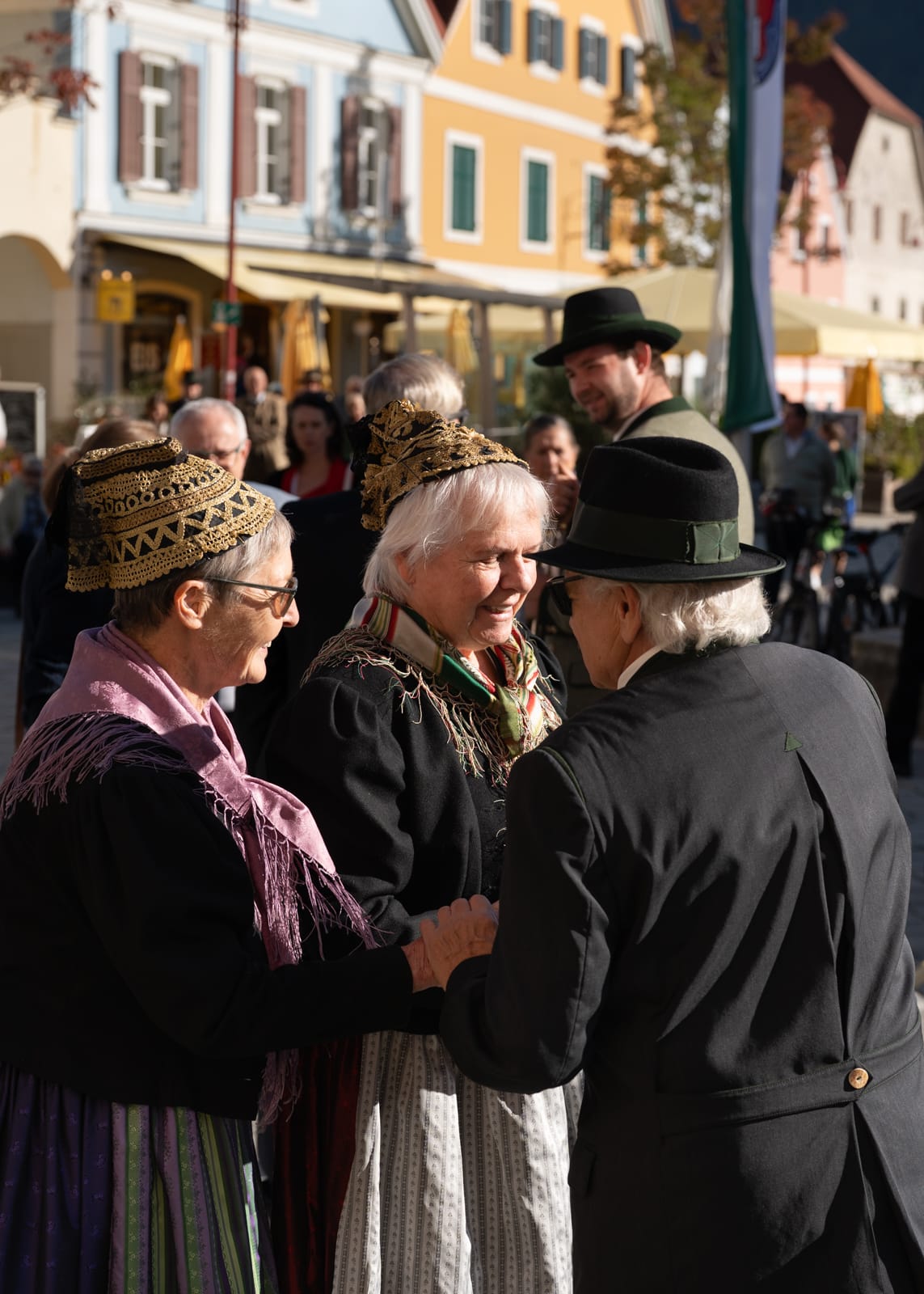 Drei Personen in Tracht auf einem Platz in Österreich mit bunten Häusern im Hintergrund.